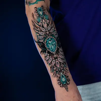 Intricate arm tattoo featuring teal gemstone and ornamental design.