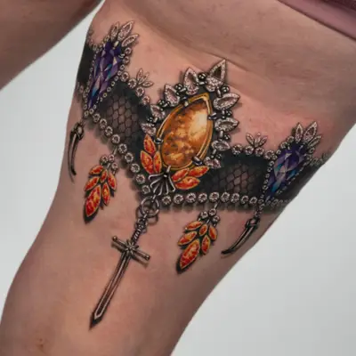 Detailed tattoo of ornamental jewelry on the back of the thigh, featuring colorful gems and a cross.