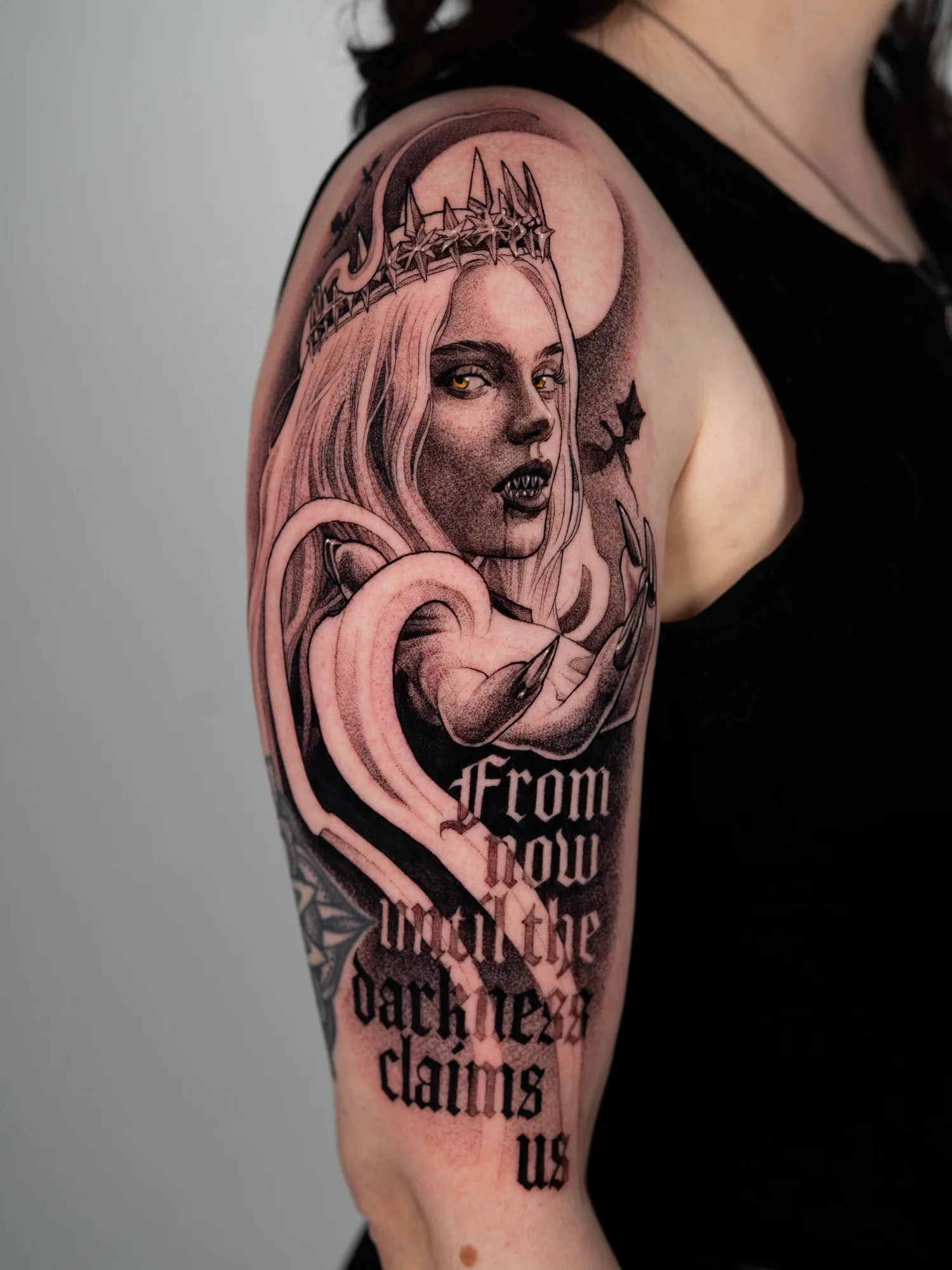 Black and grey tattoo on the upper arm of Manon Blackbeak from the Throne of Glass series in an iron crown, with gold eyes, iron-tipped nails, a full moon and bats behind her, and blackletter quote "From now until the darkness claims us"