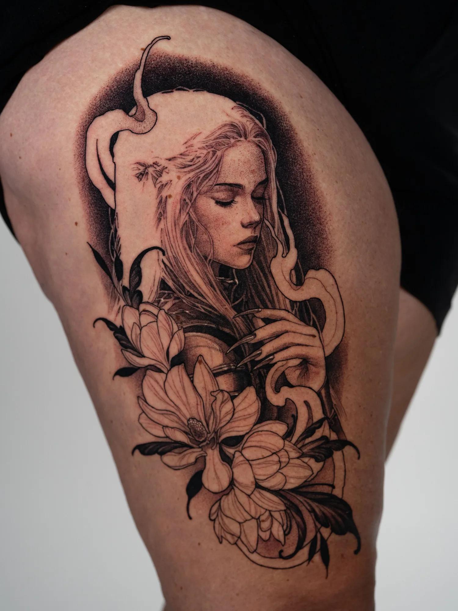 Black and grey tattoo on the right thigh of a Manon Blackbeak from the Throne of Glass series with her eyes closed and head bowed, one hand at her chest, framed by magnolias and dark ribbons of smoke.