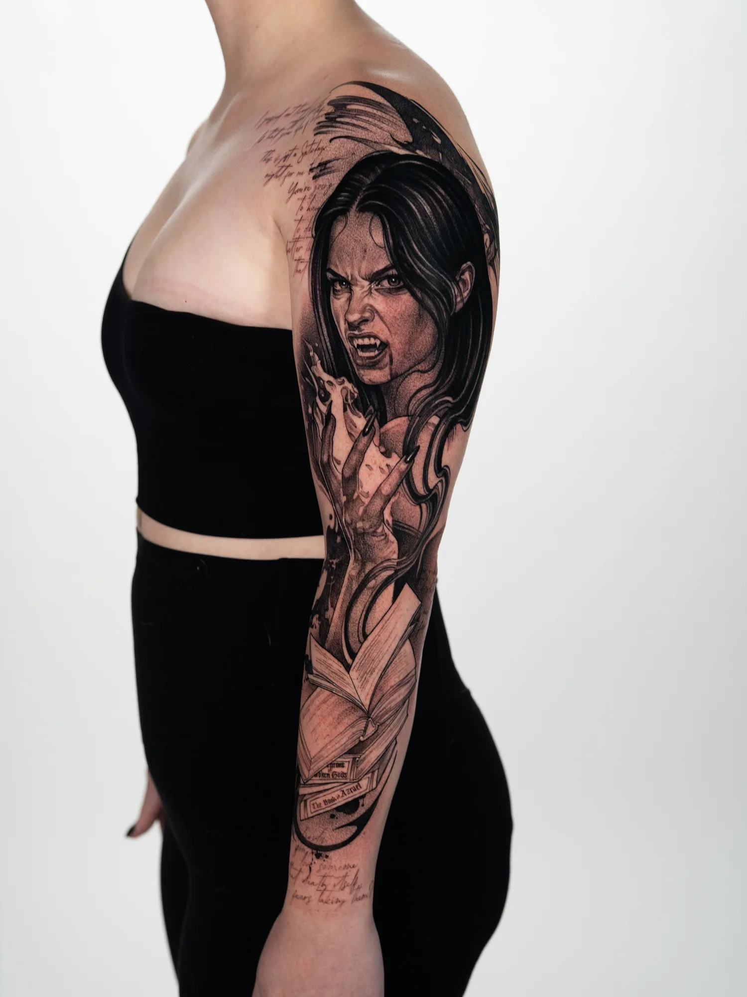Black and grey tattoo outer arm panel showing a snarling dark-haired woman with fangs holding a flame, above open books with spines reading The Book of Azrael and A Throne of Broken Gods.