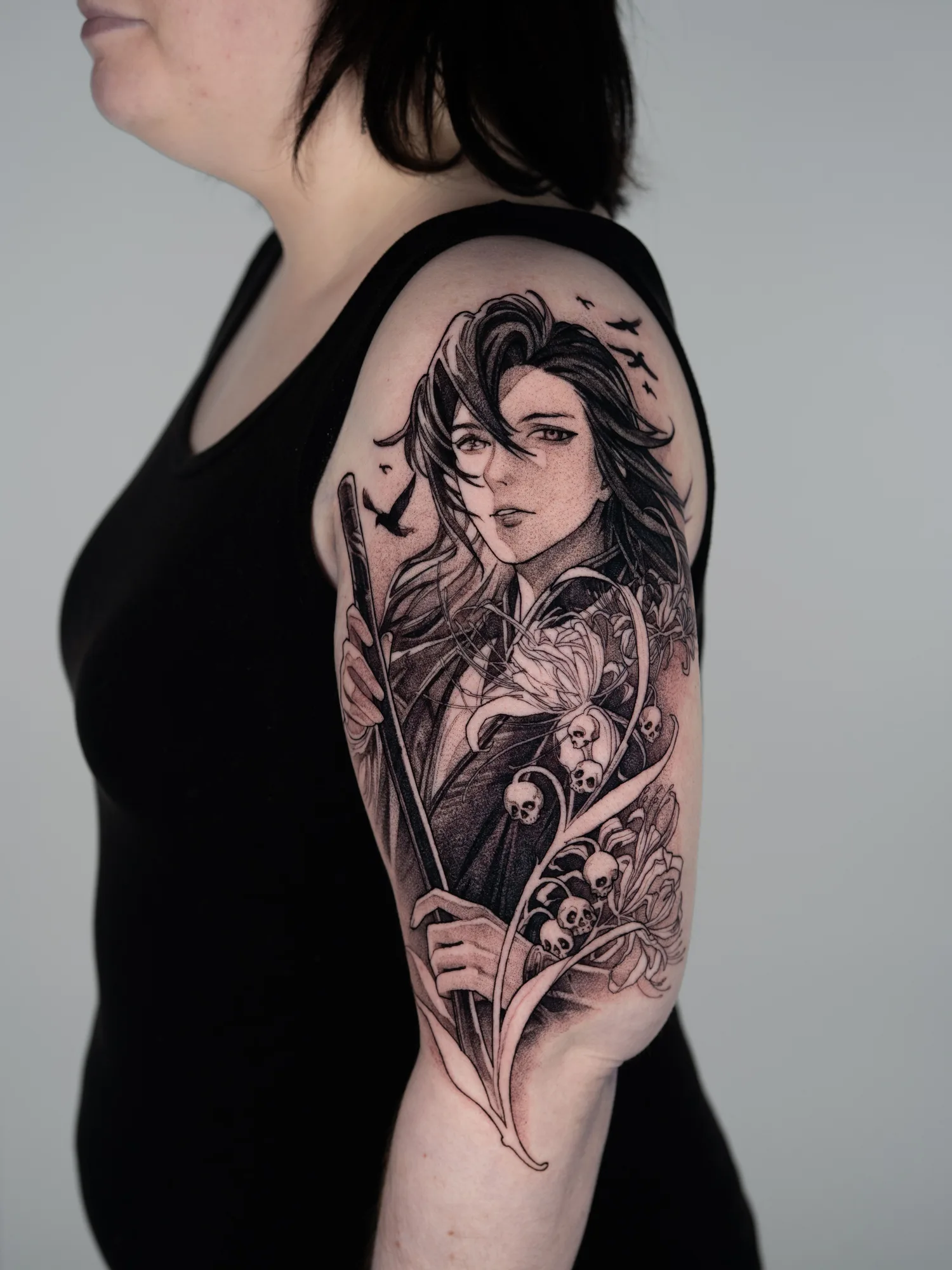 Black and grey tattoo on the outer bicep of Wei Wuxian holding a flute, surrounded by spider lilies with small skulls hanging from the stems and crows flying above.