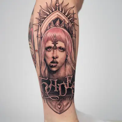 Tattoo on a lower leg of a Melanie Martinez wearing a crown, with pink hair and a third eye, set inside a gothic arch with spires and a radiating sun behind the crown, with ornate lettering across a panel below.