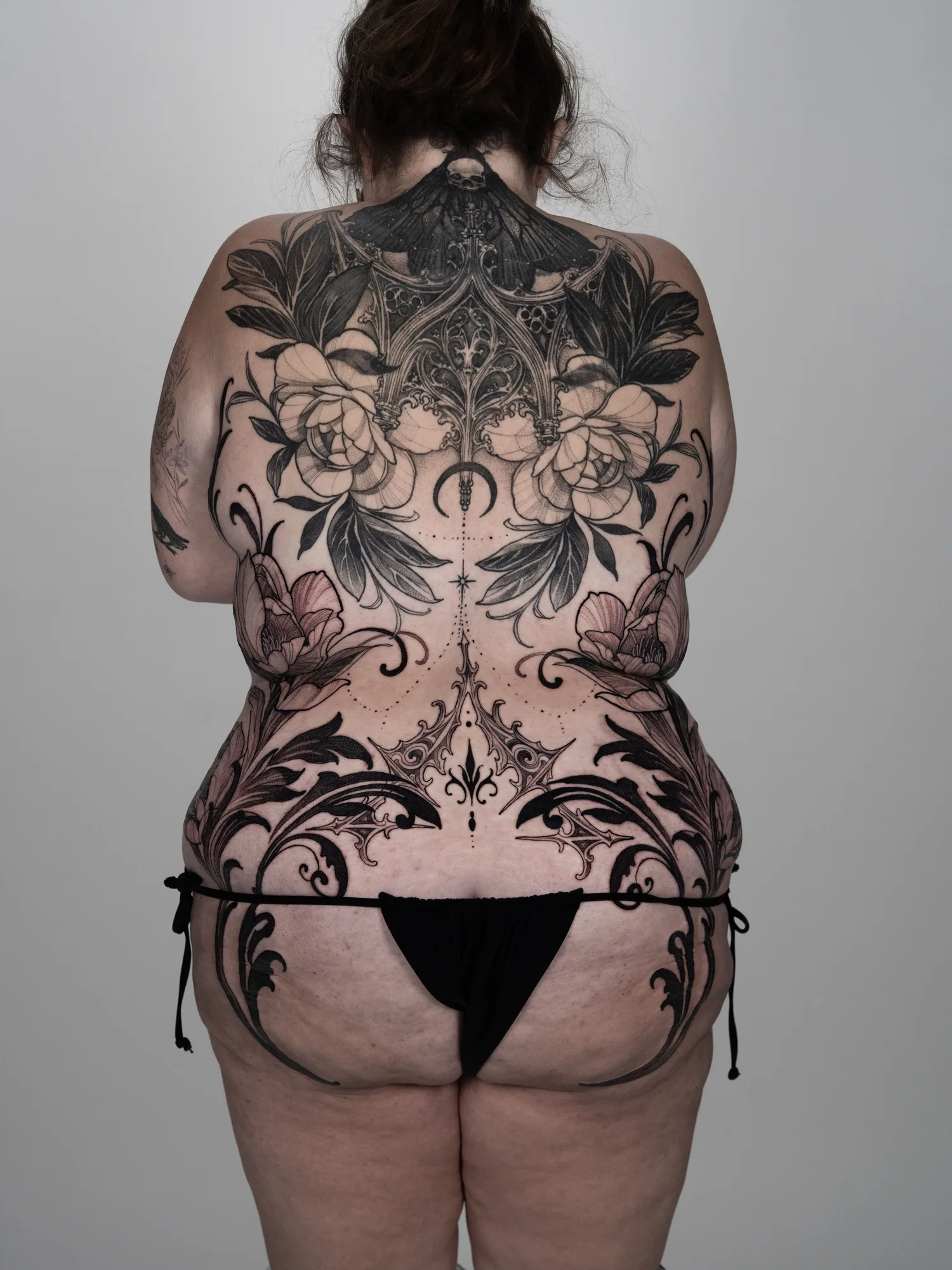 Black and grey tattoo across the entire back and both hips, showing symmetrical gothic filigree and thorned scrollwork with wild florals, meeting an existing upper back piece of a moth, gothic architecture and peonies.