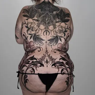 Black and grey tattoo across the entire back and both hips, showing symmetrical gothic filigree and thorned scrollwork with wild florals, meeting an existing upper back piece of a moth, gothic architecture and peonies.
