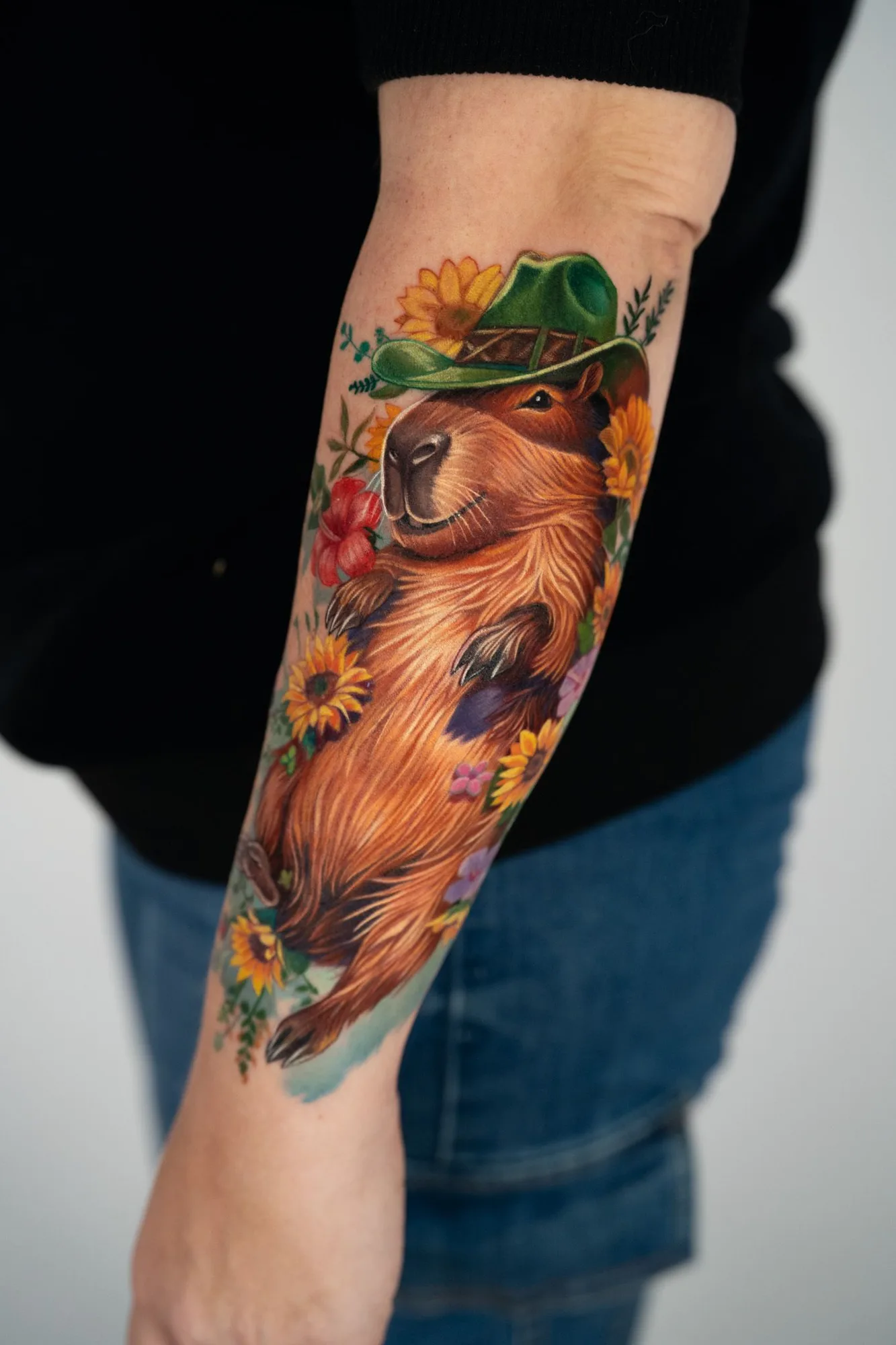 A colorful tattoo of a capybara wearing a green hat, surrounded by flowers.