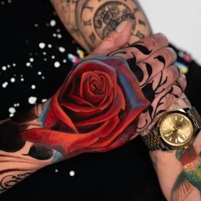 Vivid rose tattoo in red and blue on a hand, contrasted with black linework.
