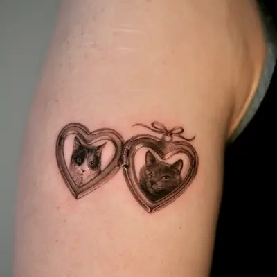 Tattoo of two cat portraits in heart-shaped lockets on upper arm.