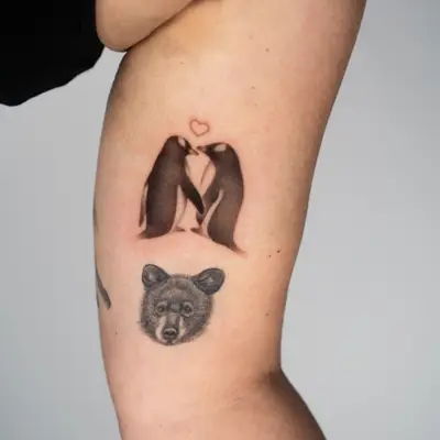Tattoo of two penguins forming a heart and a bear face on arm.