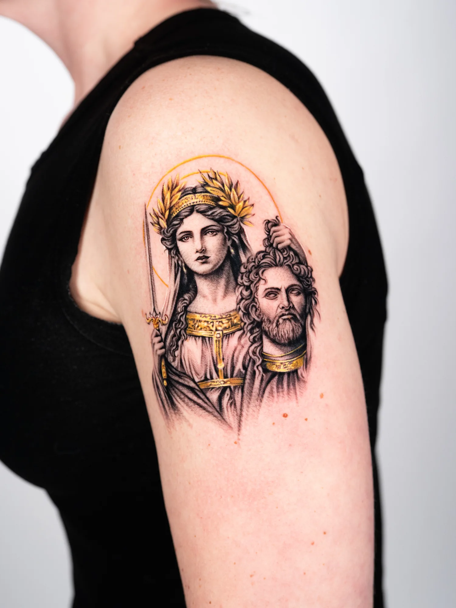 Tattoo of biblical figures on upper arm with laurel wreath and gold accents.