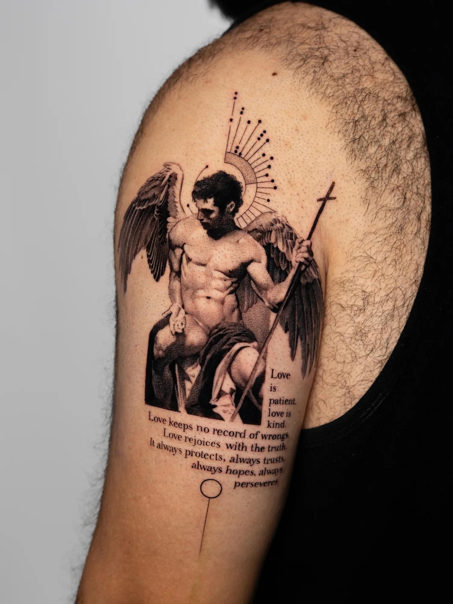 Tattoo of a seated angel in a realism and fine line style with a halo and staff, text below it.