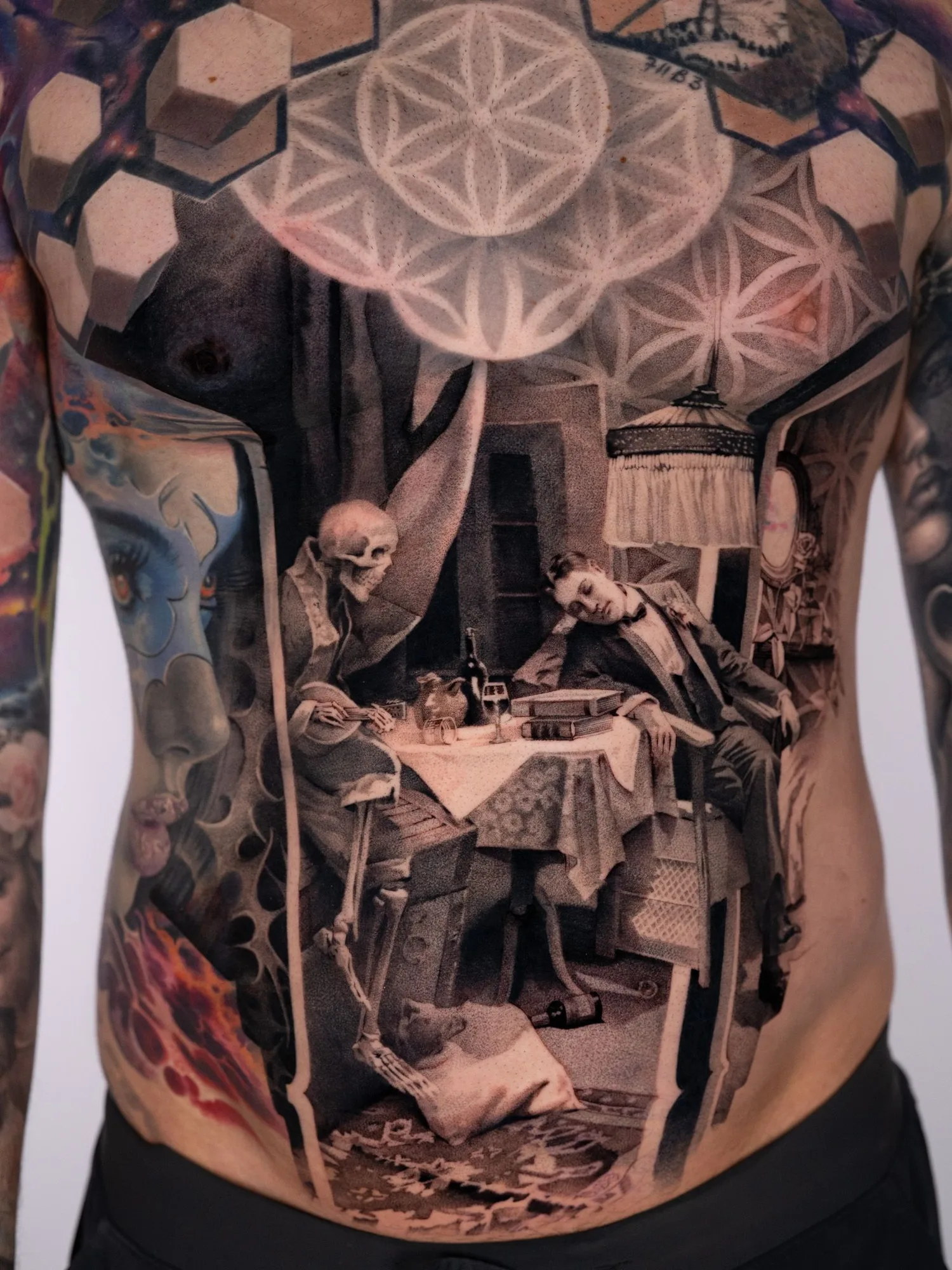 Black and grey stipple tattoo across a stomach showing a skeleton seated at a dinner table with a man in a tuxedo asleep in his chair beside it, a fringed lamp above, framed by a curtain and the sacred geometry pattern across the chest.