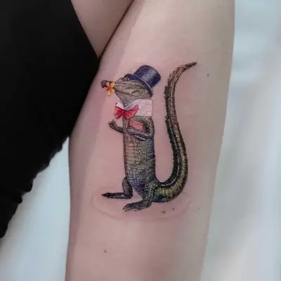 Tattoo of an alligator wearing a top hat and bow tie holding a flower.