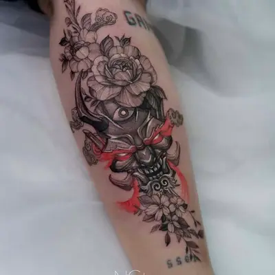 Tattoo of a demon mask with floral and watercolor elements on forearm.