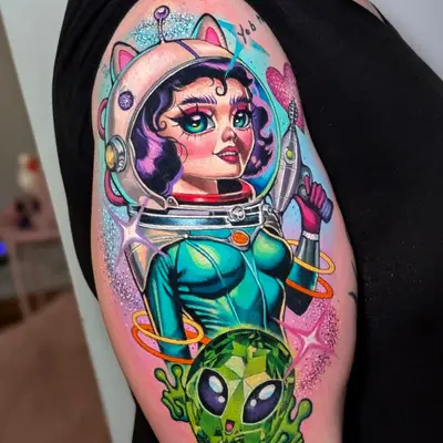 Color tattoo on the outer upper arm of a retro space girl in a cat-eared bubble helmet holding a ray gun, with a green gemstone alien below her, orange rings, sparkles and a glitter heart.