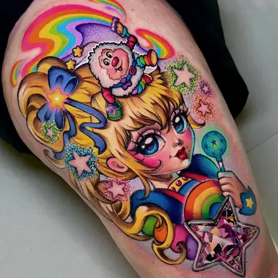 Color tattoo above the knee of Rainbow Brite and her sprite Twink, with a rainbow trail, glitter stars and a faceted pink star-shaped jewel on her wand, in an illustrative pop art style.