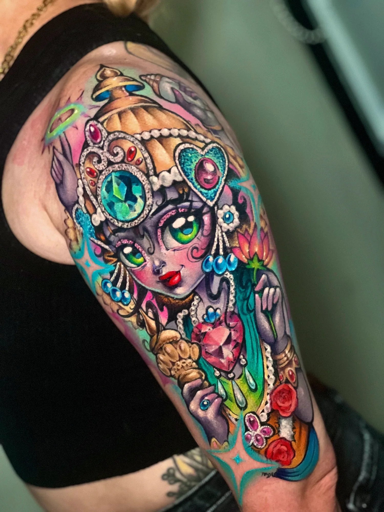 Color tattoo on the outer upper arm of a four-armed goddess in a doll-like style, wearing a jeweled gold crown and pearls and holding a pink lotus, a conch shell and a golden mace, with glitter gems and red roses.
