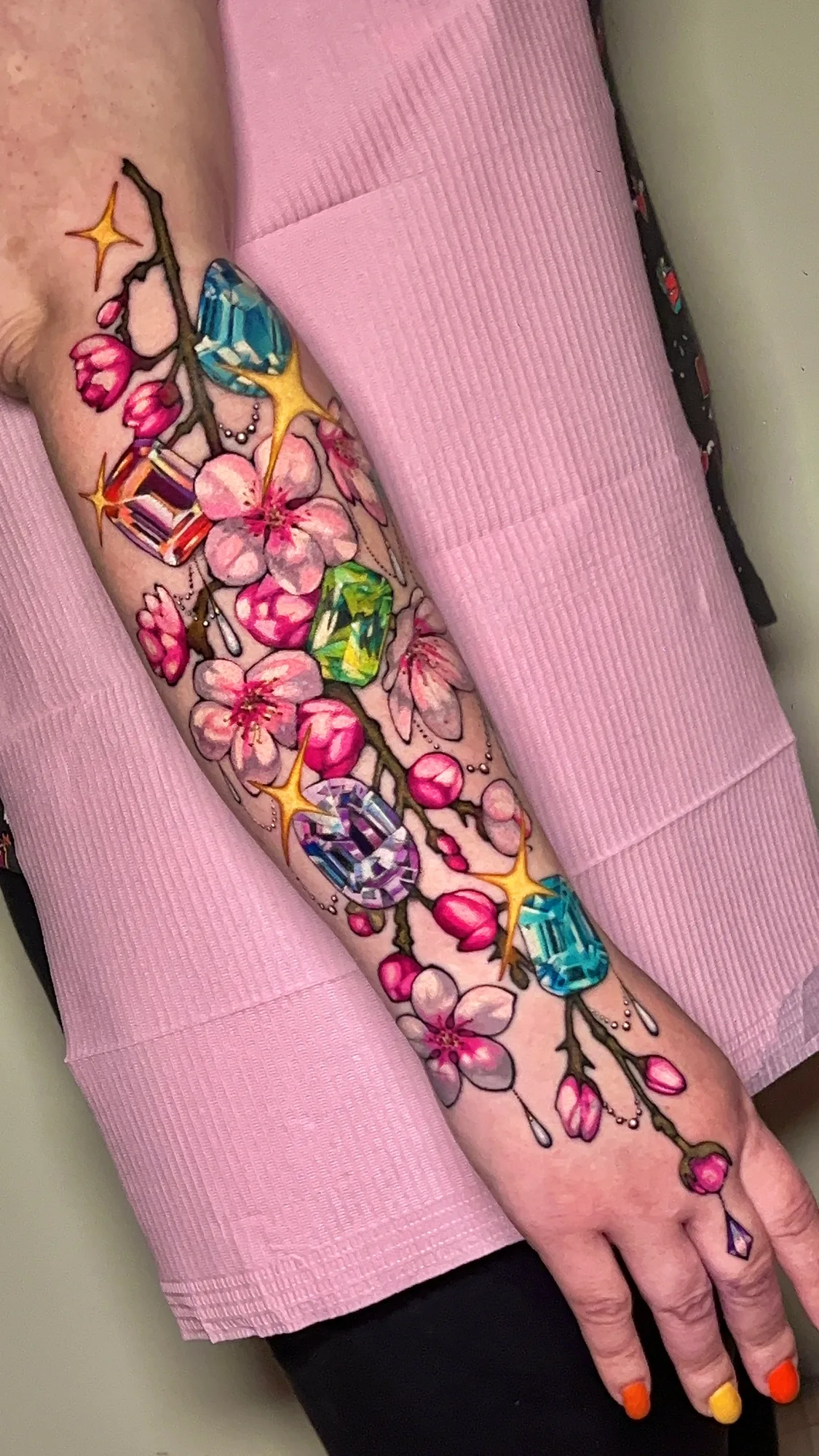 Color tattoo of a cherry blossom branch running from the forearm across the wrist onto the back of the hand, set with cut gemstones in blue, amber, green and lavender and gold sparkles
