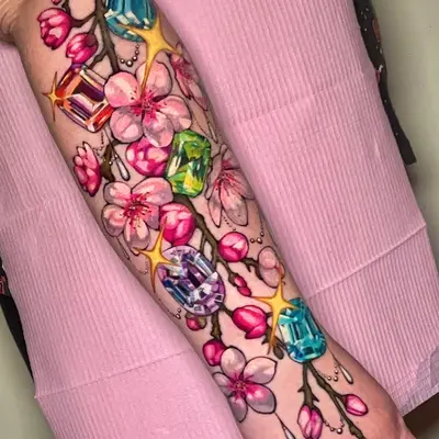 Color tattoo of a cherry blossom branch running from the forearm across the wrist onto the back of the hand, set with cut gemstones in blue, amber, green and lavender and gold sparkles