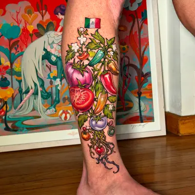 Color tattoo on the shin of a tomato and pepper plant growing from tangled roots at the ankle, with purple, red and blue tomatoes, red, green and orange chiles, white blossoms and a small Mexican flag at the top.