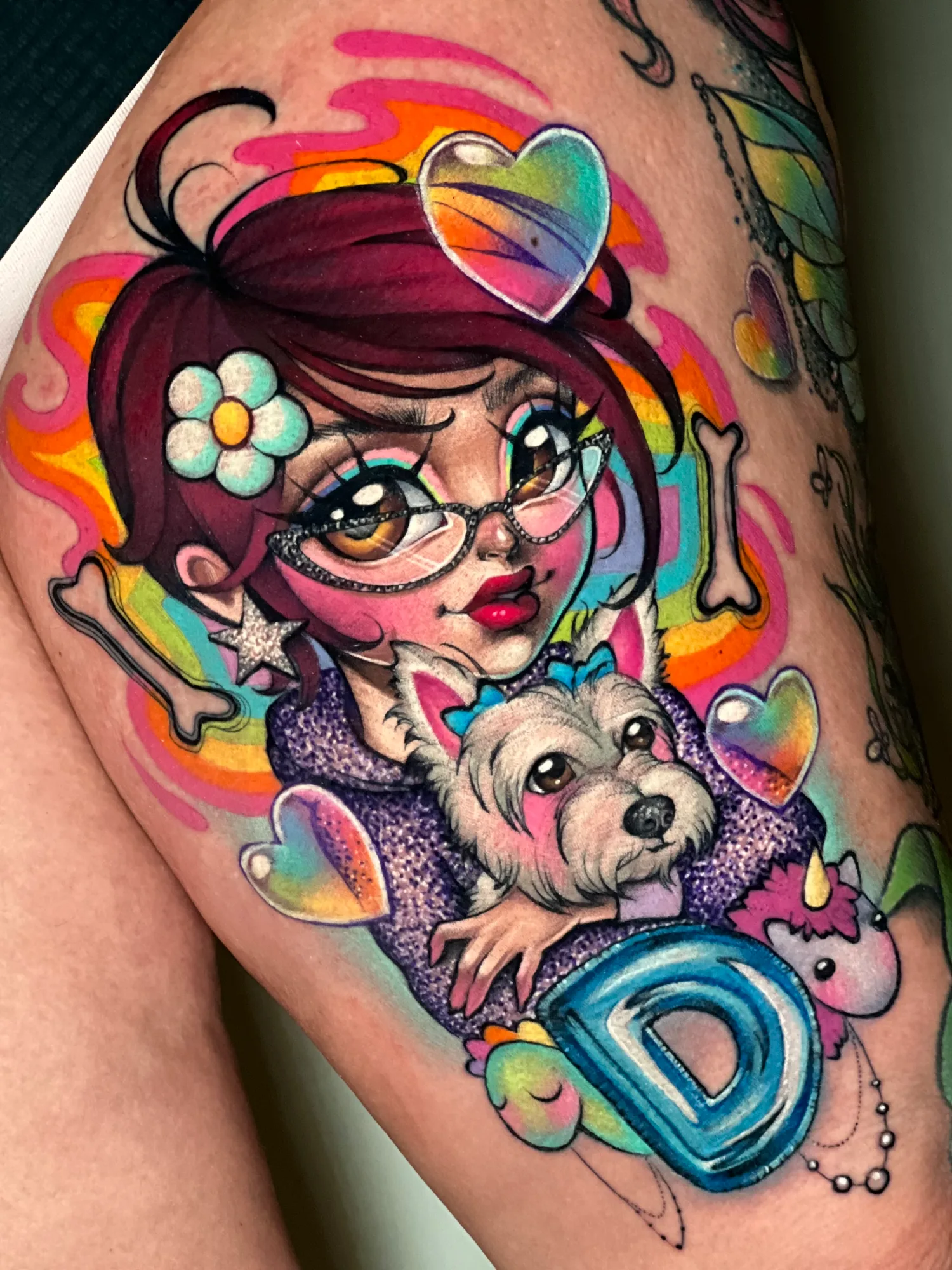 Color tattoo on a thigh of a cartoon girl in cat-eye glasses holding a small white dog, with glossy hearts, bones, a plush unicorn and a blue letter D around them on a rainbow background