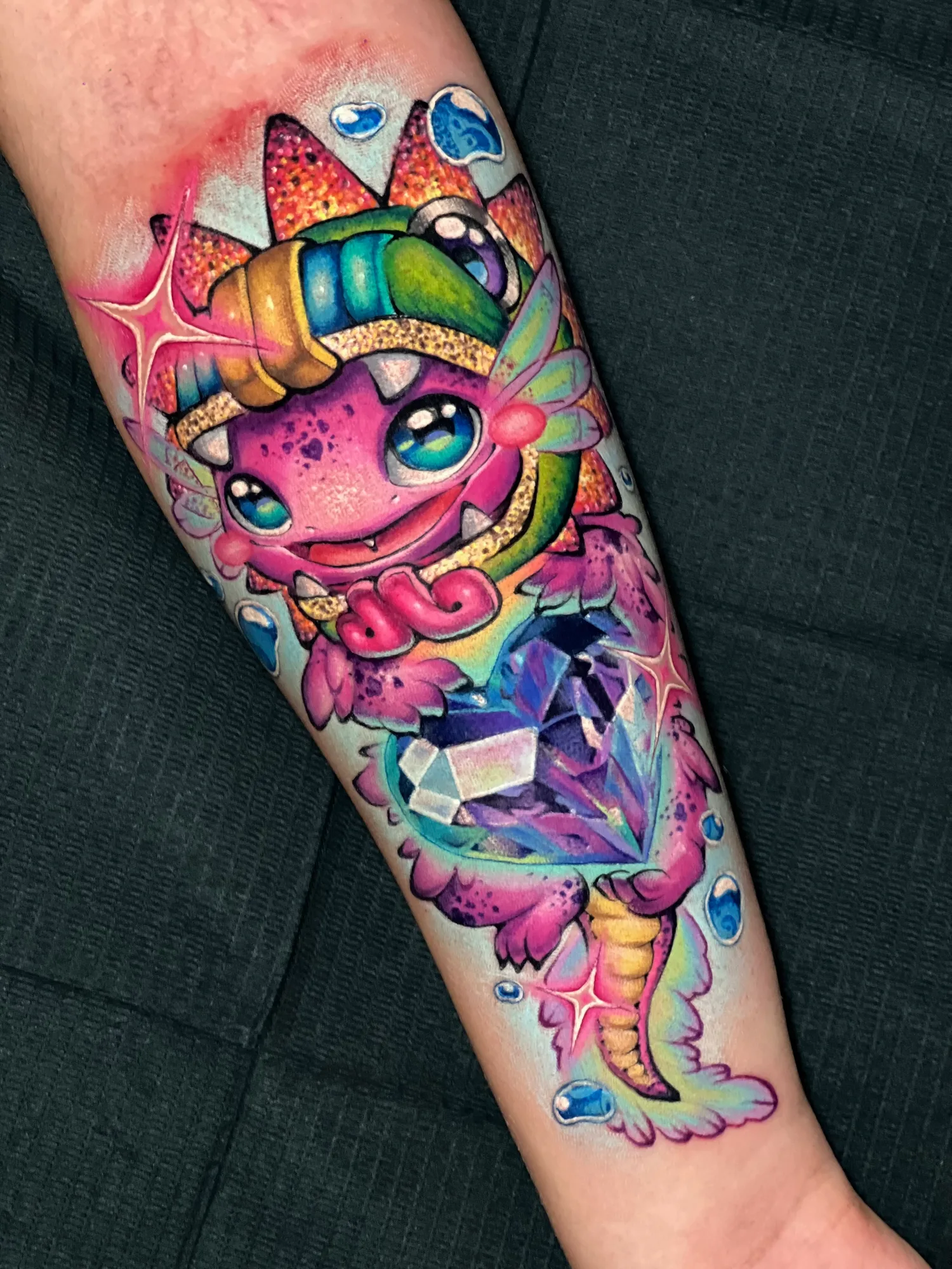 Color tattoo on a forearm of a cute pink baby dragon in a striped headband hugging a large faceted purple gem, with sparkles and blue water droplets on a pale blue glow