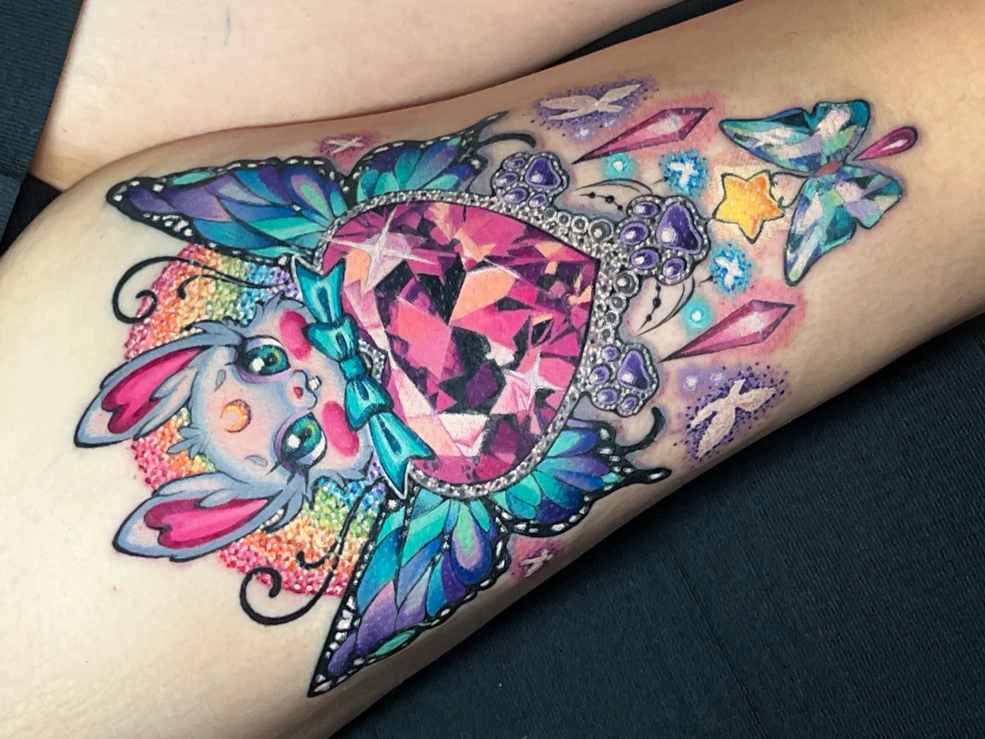 Color tattoo on a thigh of a cute grey bunny with heart-shaped ears holding a faceted pink heart gem with butterfly wings, a rainbow glitter halo behind it and crystals, paw-print jewels and a small glass butterfly below