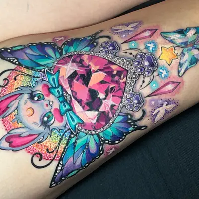 Color tattoo on a thigh of a cute grey bunny with heart-shaped ears holding a faceted pink heart gem with butterfly wings, a rainbow glitter halo behind it and crystals, paw-print jewels and a small glass butterfly below