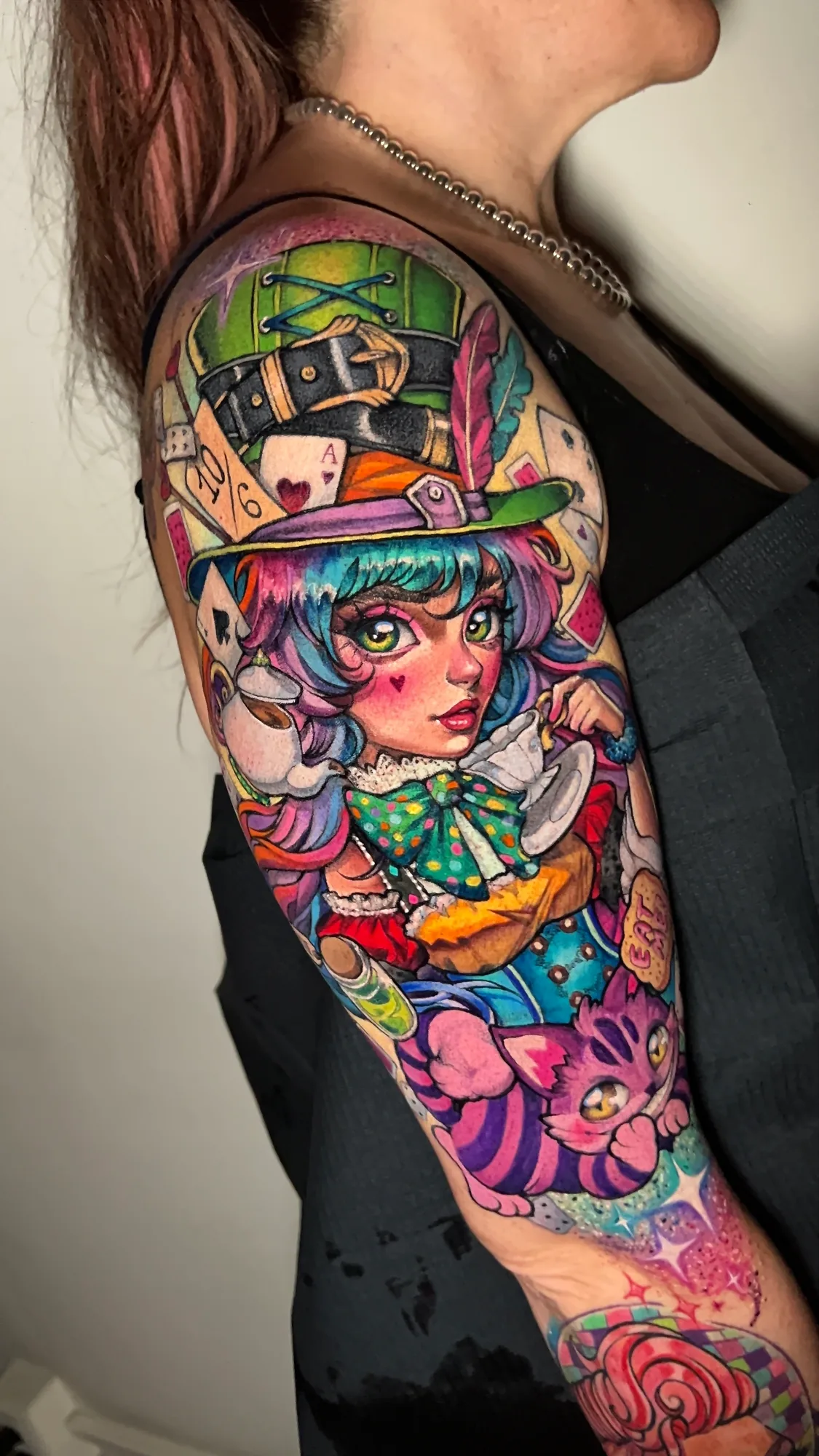 Color half sleeve tattoo on an upper arm of an anime-style girl as the Mad Hatter in a tall green top hat, sipping from a teacup, with playing cards behind her and the Cheshire Cat below