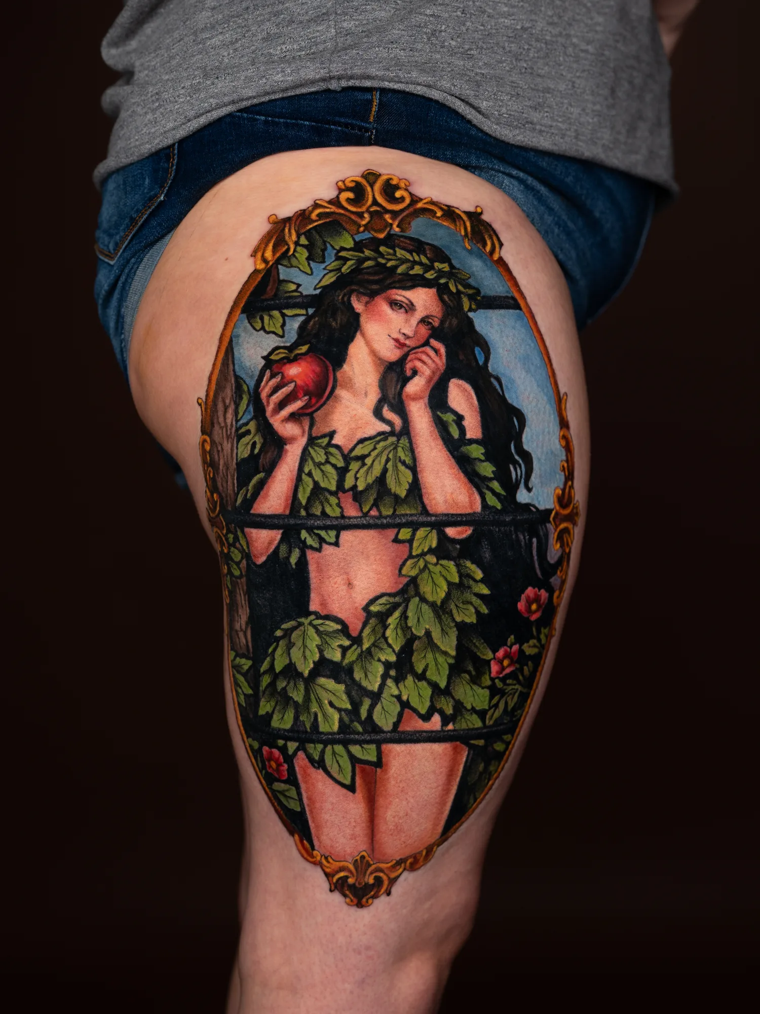 Tattoo of a woman in foliage with an apple on thigh.