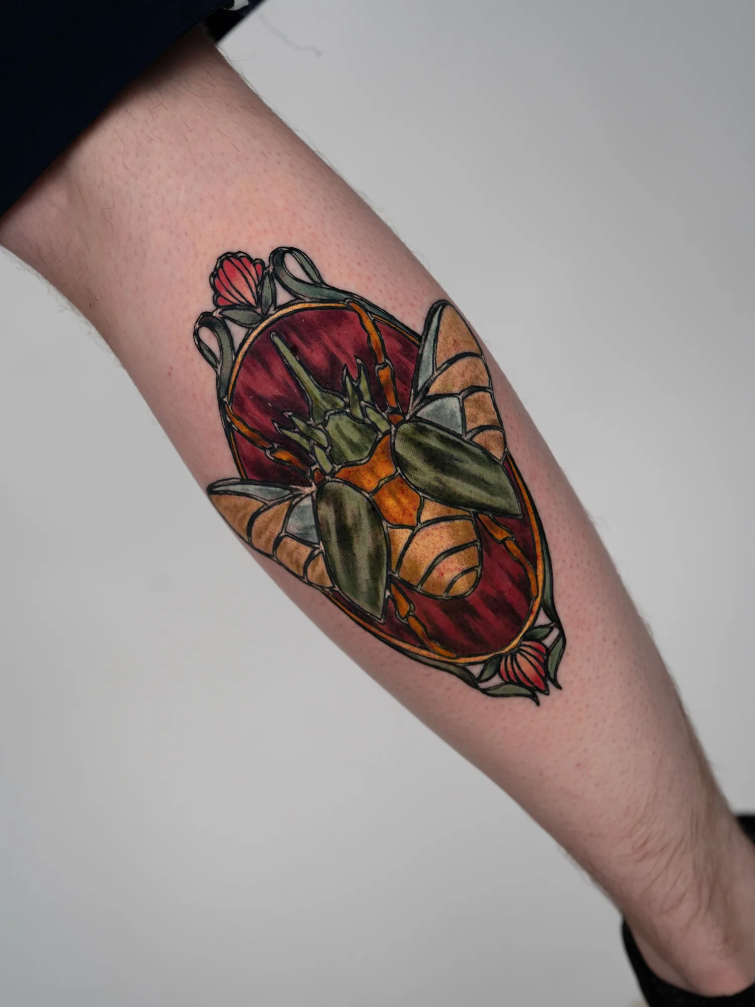 Color stained glass style tattoo of a beetle with green wing cases inside an amber and red oval with tulips, on the back of a calf.