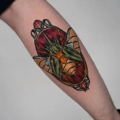 Color stained glass style tattoo of a beetle with green wing cases inside an amber and red oval with tulips, on the back of a calf.