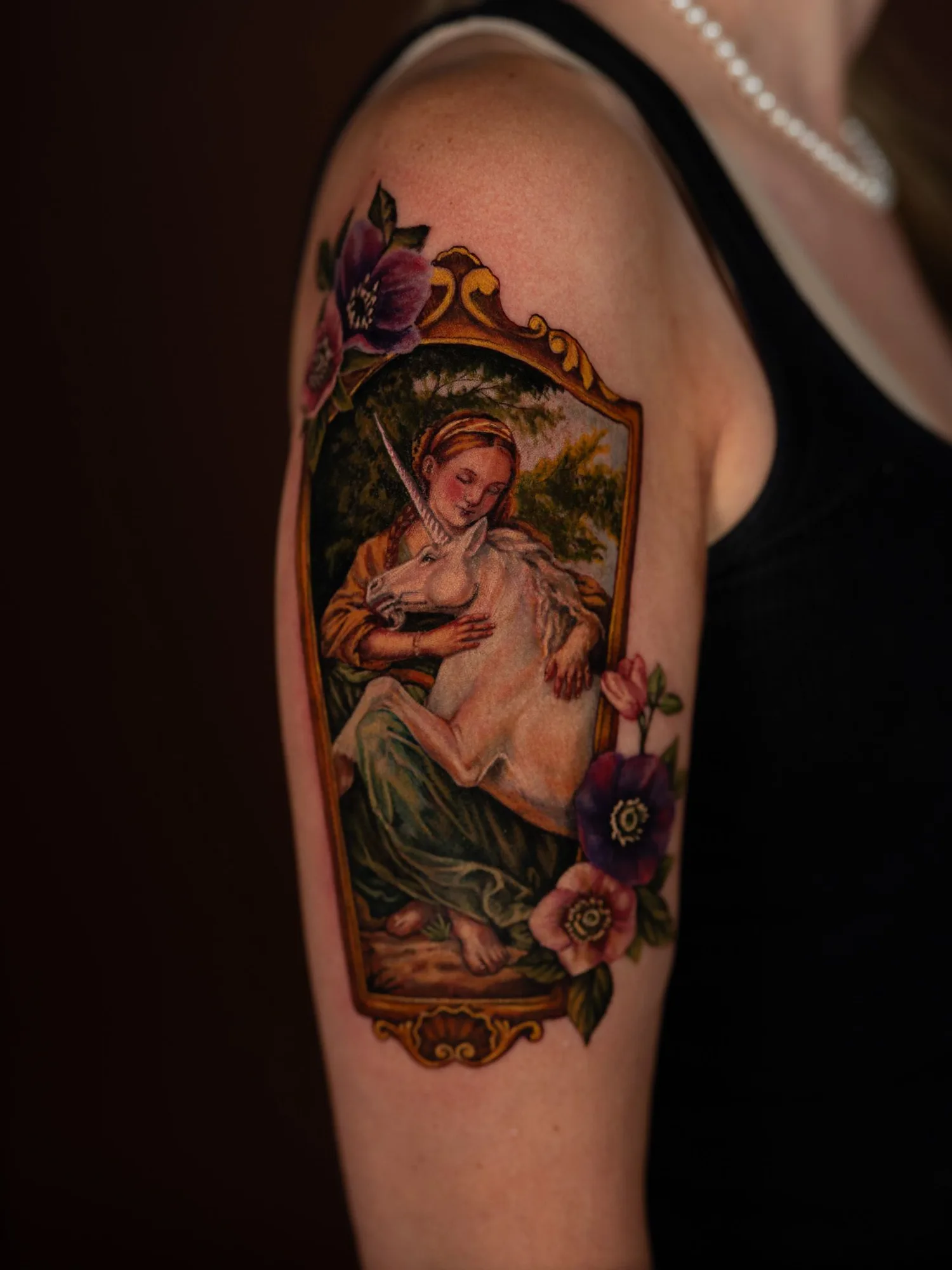 Color painterly tattoo of a woman in a gold frame with her arms around a white unicorn, surrounded by anemones, on an upper outer arm.
