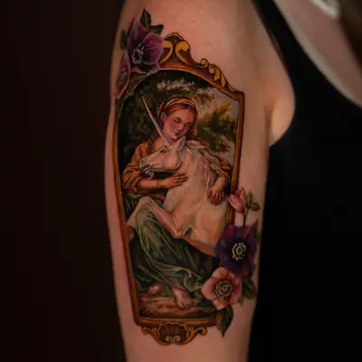 Color painterly tattoo of a woman in a gold frame with her arms around a white unicorn, surrounded by anemones, on an upper outer arm.