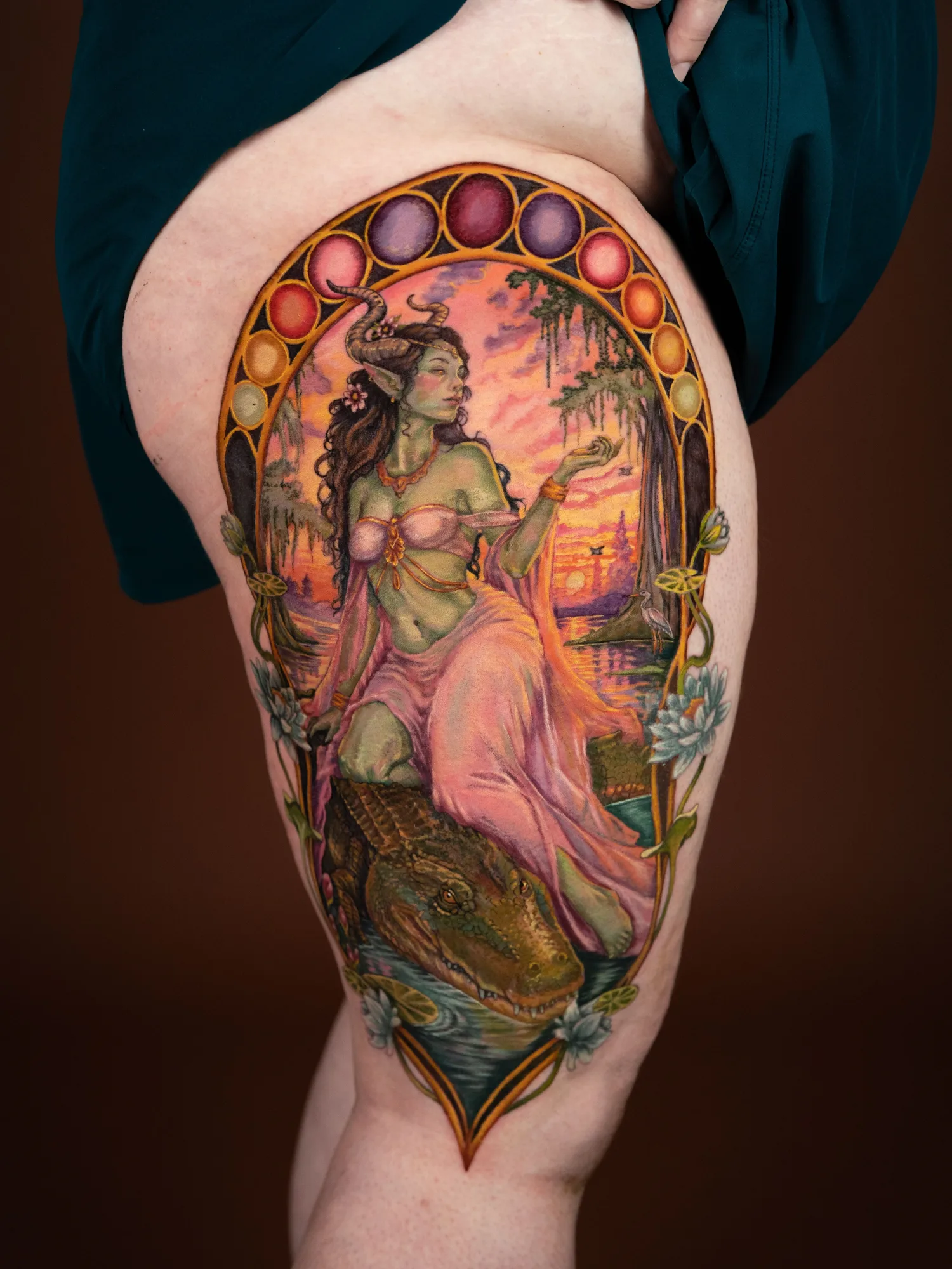 Color Art Nouveau tattoo of a green horned woman in a pink gown reclining against an alligator in a sunset bayou, inside an arched mosaic frame, on a front thigh panel.
