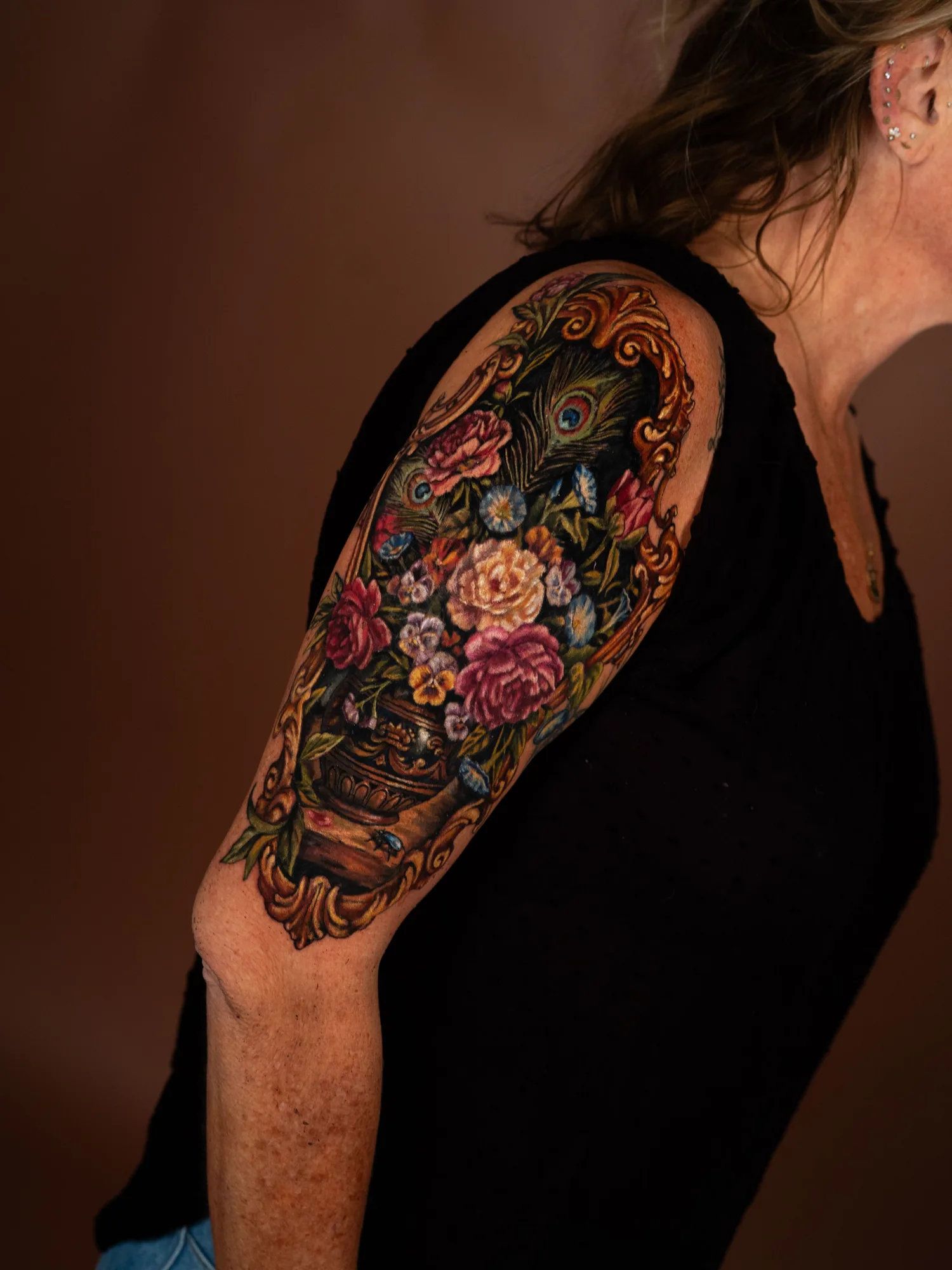 Color painterly tattoo of a Dutch still life bouquet with peonies, roses, pansies and peacock feathers in an urn, inside a carved gold frame, on an upper outer arm.