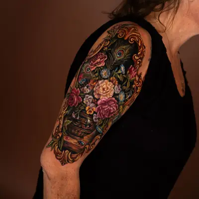 Color painterly tattoo of a Dutch still life bouquet with peonies, roses, pansies and peacock feathers in an urn, inside a carved gold frame, on an upper outer arm.