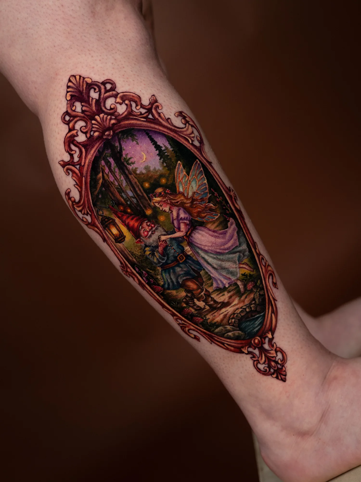 Color painterly tattoo of a gnome and a winged fairy dancing in a moonlit forest inside an ornate copper frame, on an outer calf.