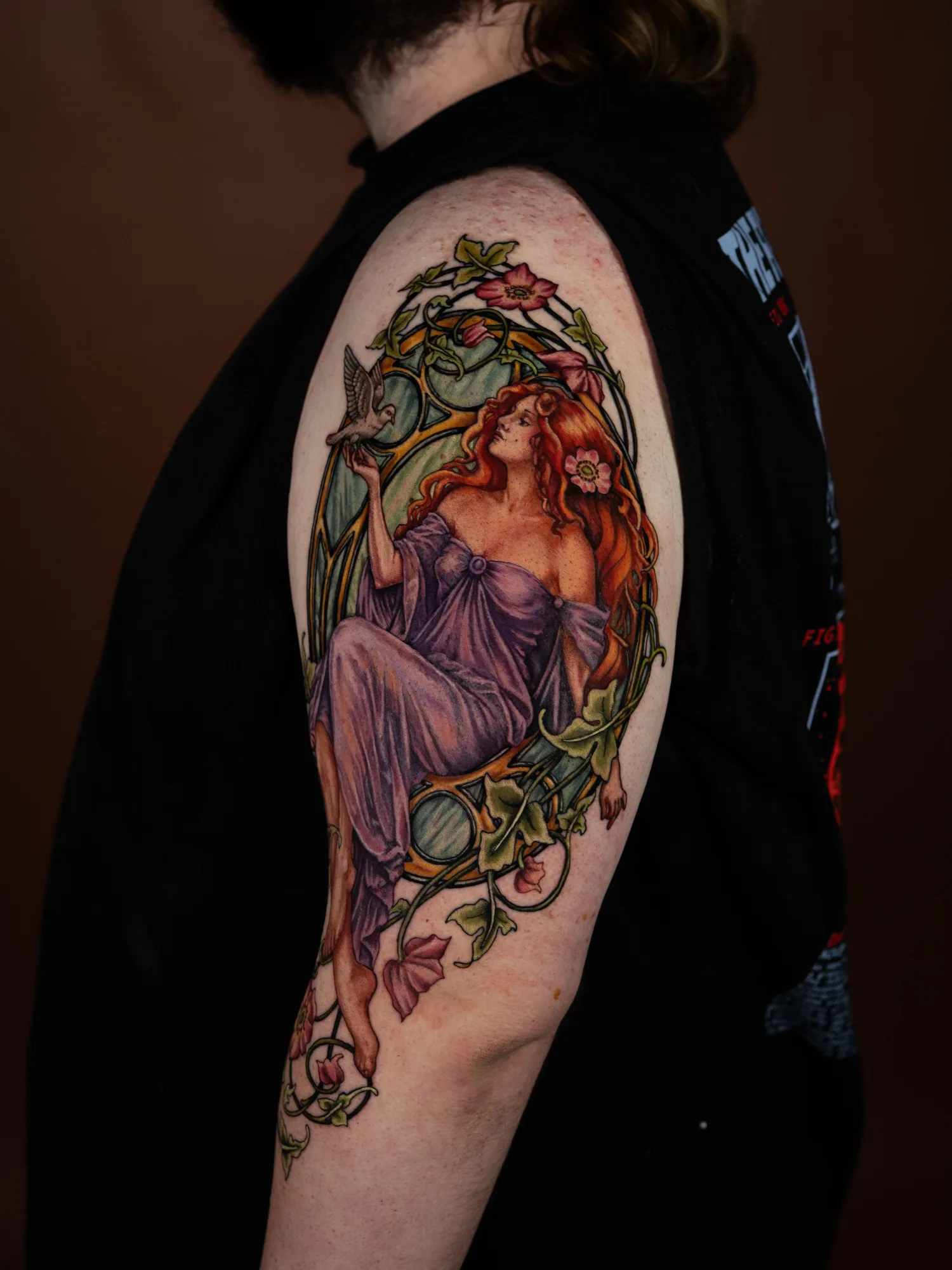 Color Art Nouveau tattoo of a red haired woman in a violet gown releasing a dove, inside a gold frame with ivy and wild roses breaking past its border, on an upper outer arm.