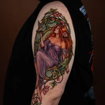 Color Art Nouveau tattoo of a red haired woman in a violet gown releasing a dove, inside a gold frame with ivy and wild roses breaking past its border, on an upper outer arm.