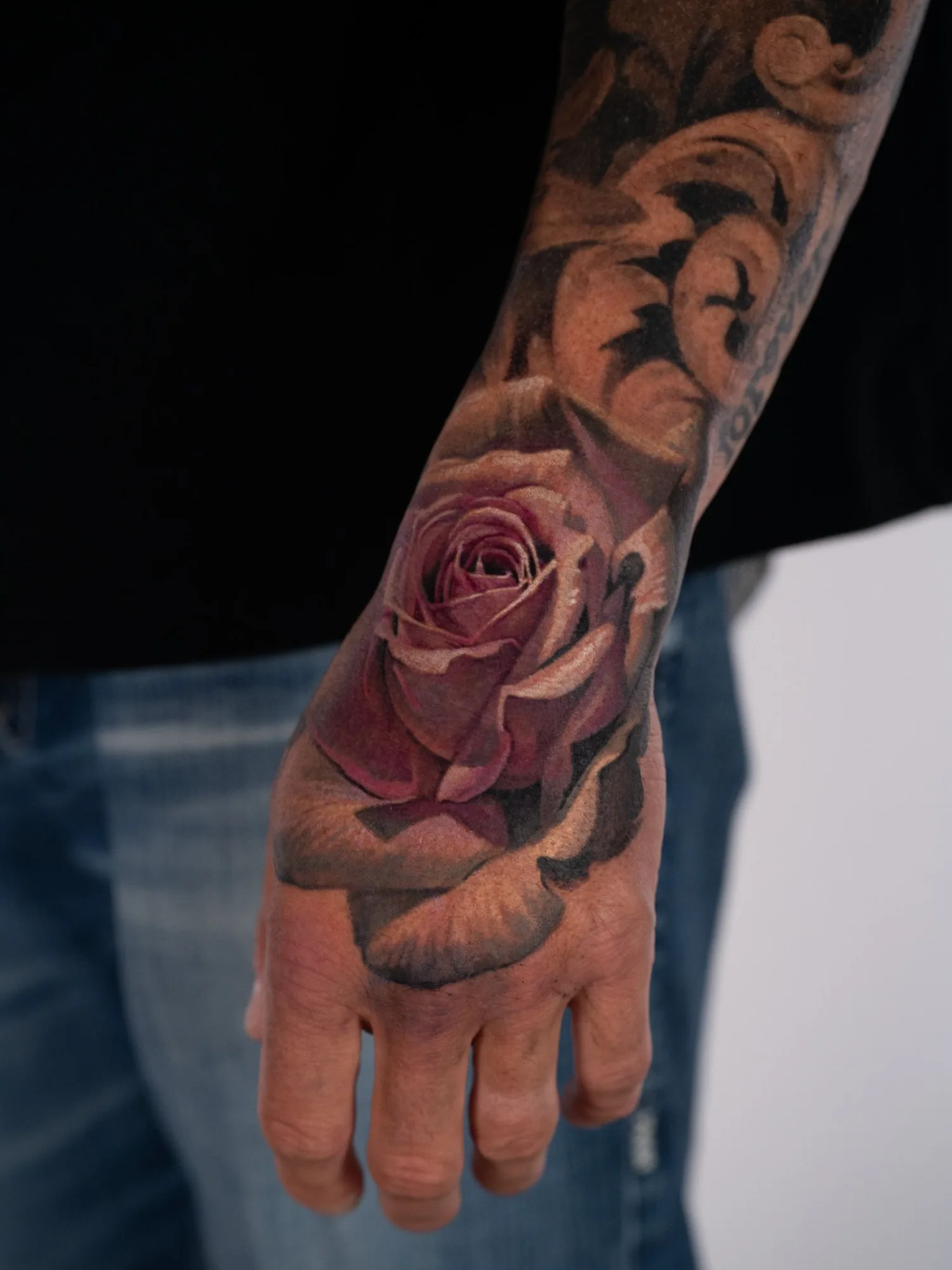 Color realism tattoo of a single dusty pink rose on the back of a hand, worked into an existing black and grey filigree sleeve.