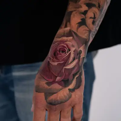 Color realism tattoo of a single dusty pink rose on the back of a hand, worked into an existing black and grey filigree sleeve.