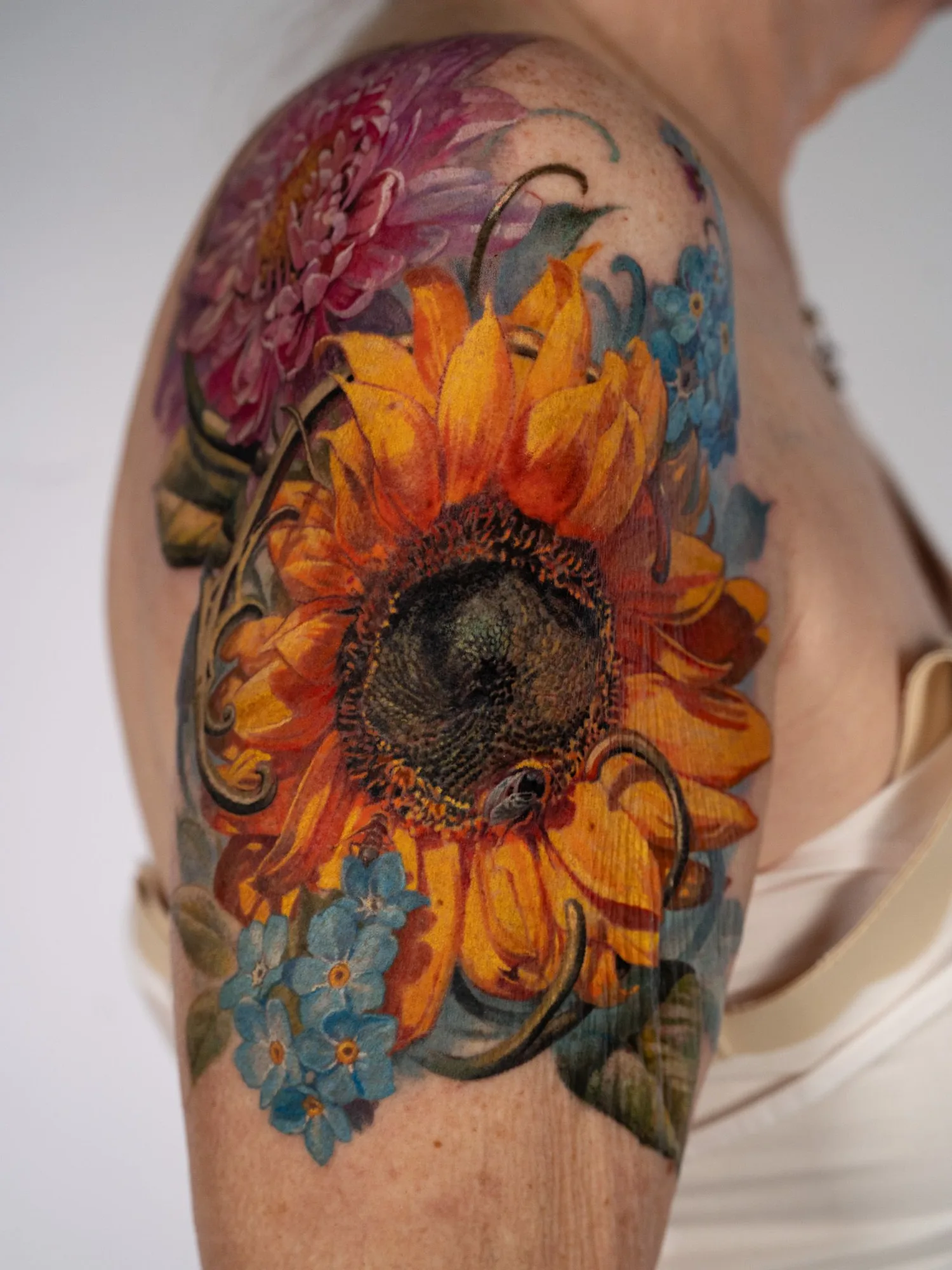 Color realism tattoo of a large sunflower with a bee, a magenta chrysanthemum and forget-me-nots, wrapping a shoulder cap.