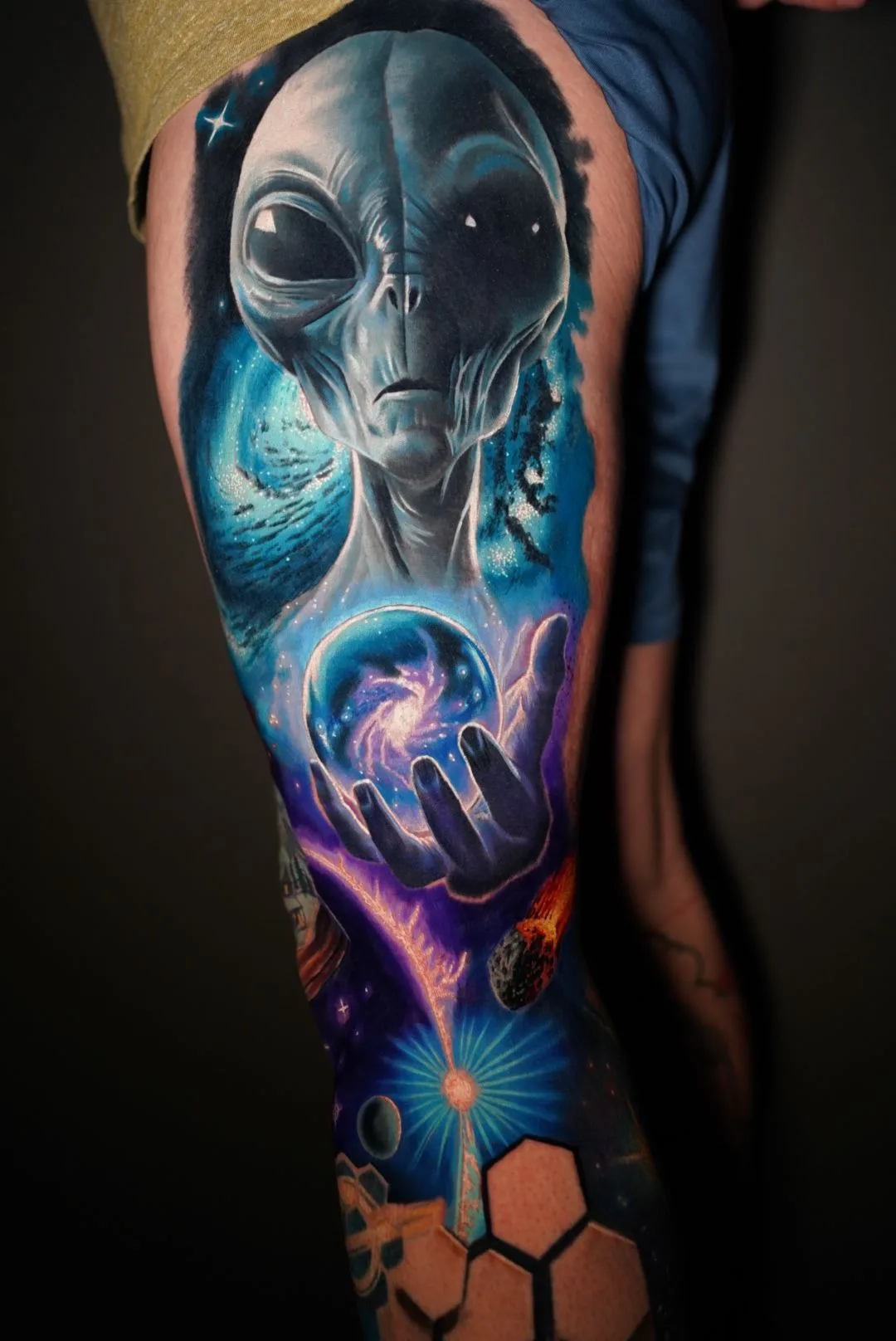 Tattoo of an alien in a surrealism style holding an orb with a galaxy on an upper thigh.