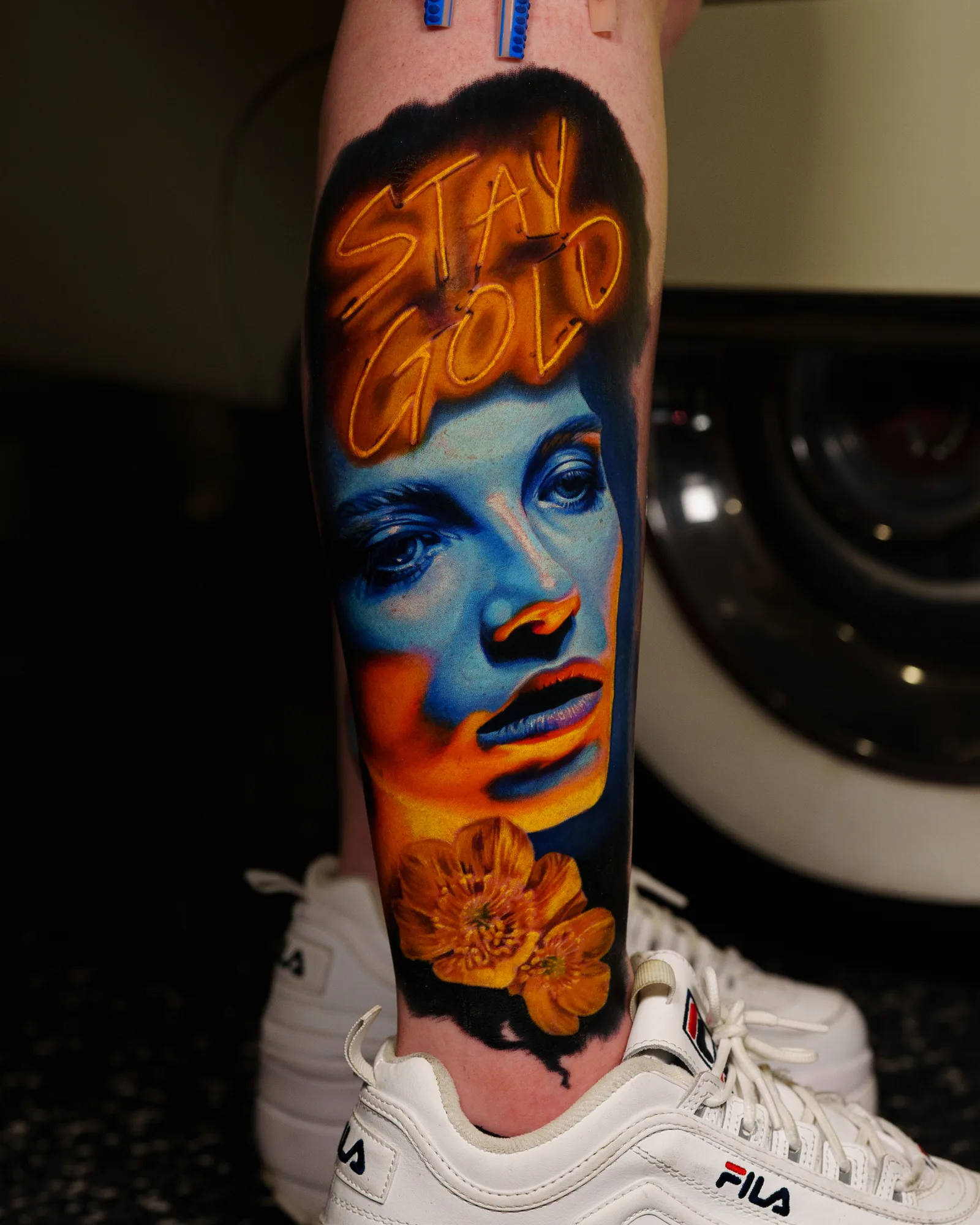 Tattoo of a woman's face with 'Stay Gold' text and flowers, in vivid colors.