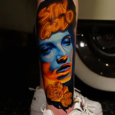 Tattoo of a woman's face with 'Stay Gold' text and flowers, in vivid colors.