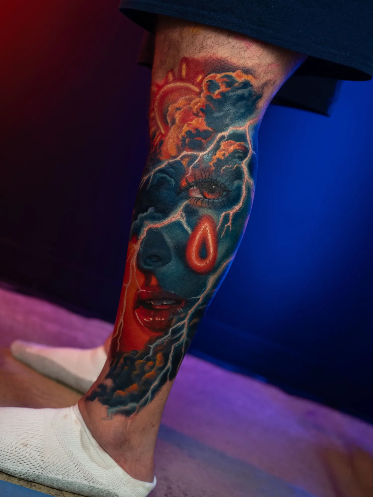 A vibrant leg tattoo featuring a face, raindrops, and storm clouds in vivid colors with an orange and blue neon palette.
