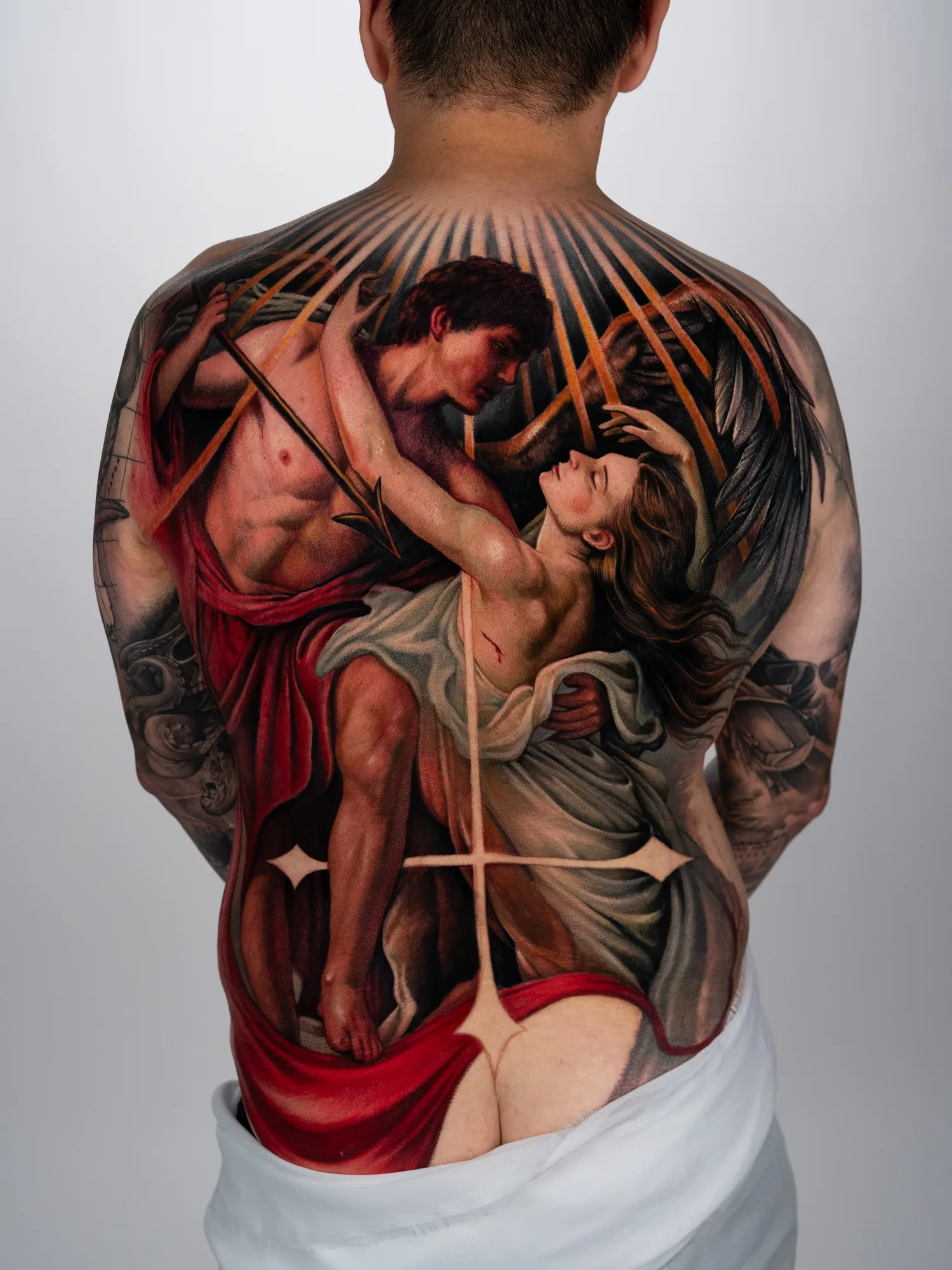 Tattoo depicting an embracing couple with draped fabrics and rays on a back piece.