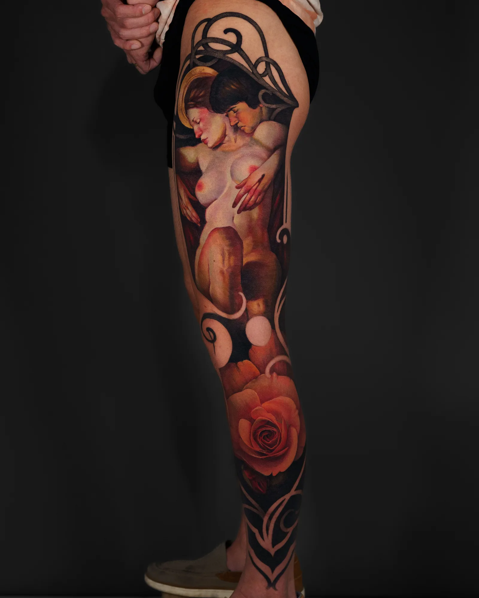 Tattoo on thigh featuring two nude figures with flowers and ornate elements.