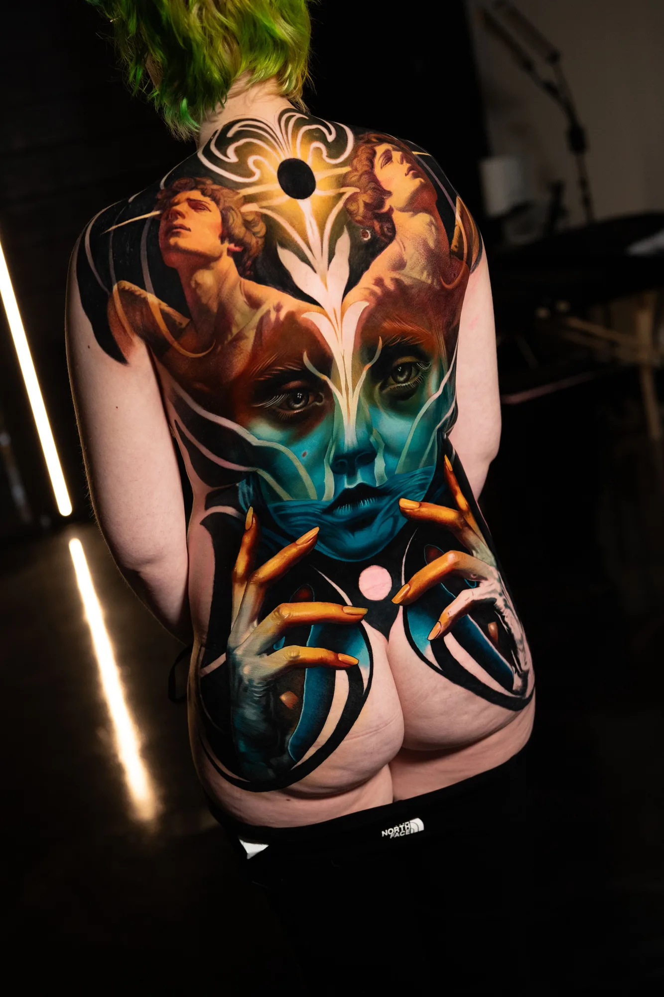Full back piece tattoo done in a surrealism style with color, a portrait, hands, and women.